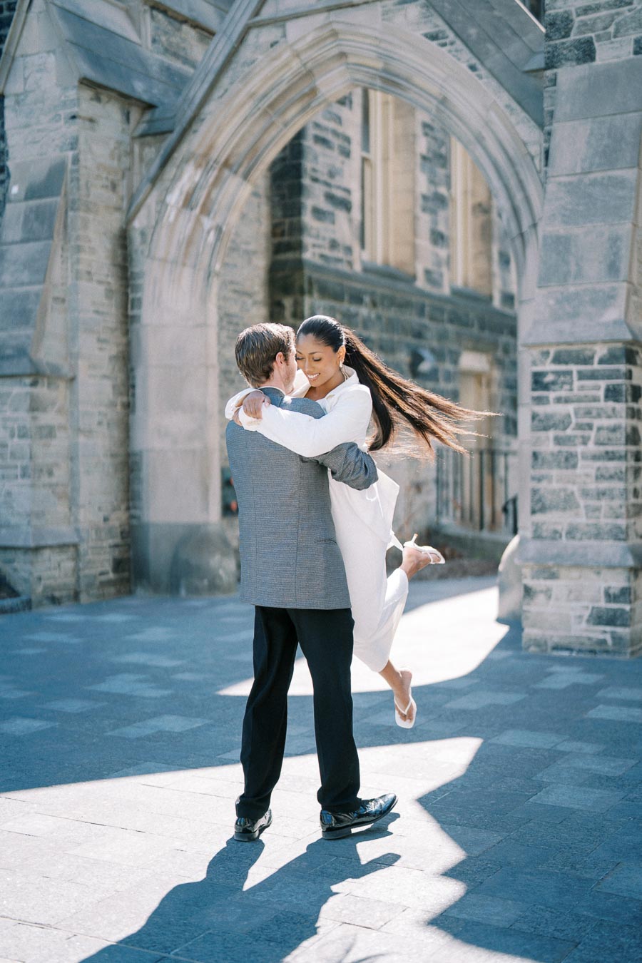 Couple embracing joyfully under historic stone archway on a sunny day.