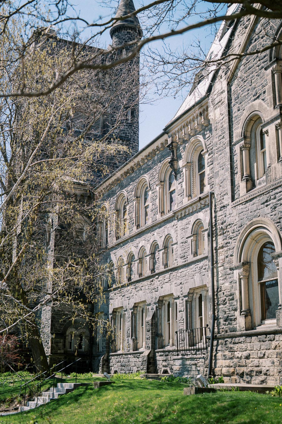 Historic stone building with arched windows and tower, under a clear blue sky, surrounded by lush green lawn.