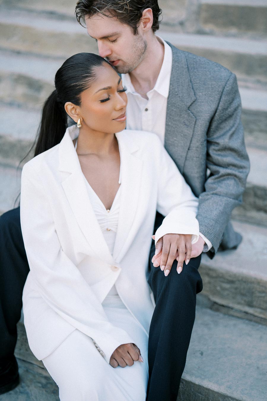 Couple seated on steps, woman in white suit holding a man's hand, with a loving and serene expression.