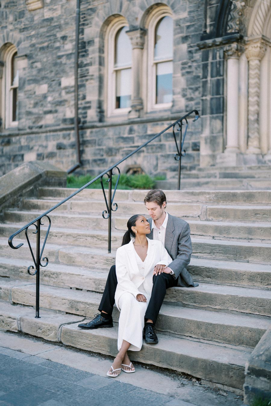 Couple sitting on stone steps of historic building, sharing a tender moment in stylish formal attire.
