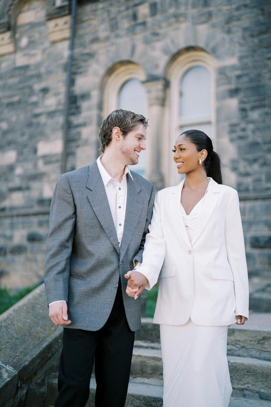 Couple in formal attire smiling and holding hands on stone steps, with a historic building in the background.