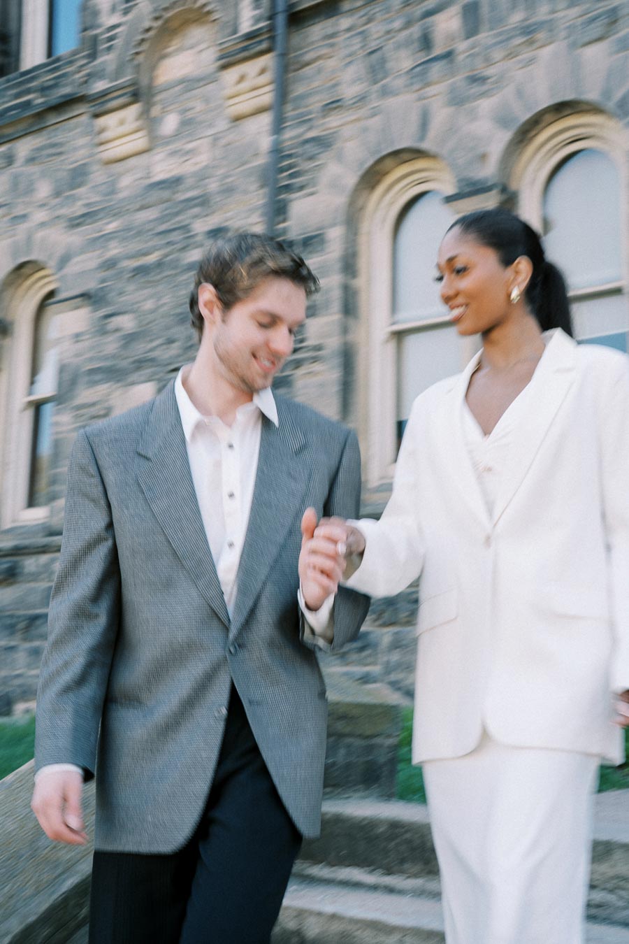 Two people in formal attire holding hands while walking down stone steps outside a building.