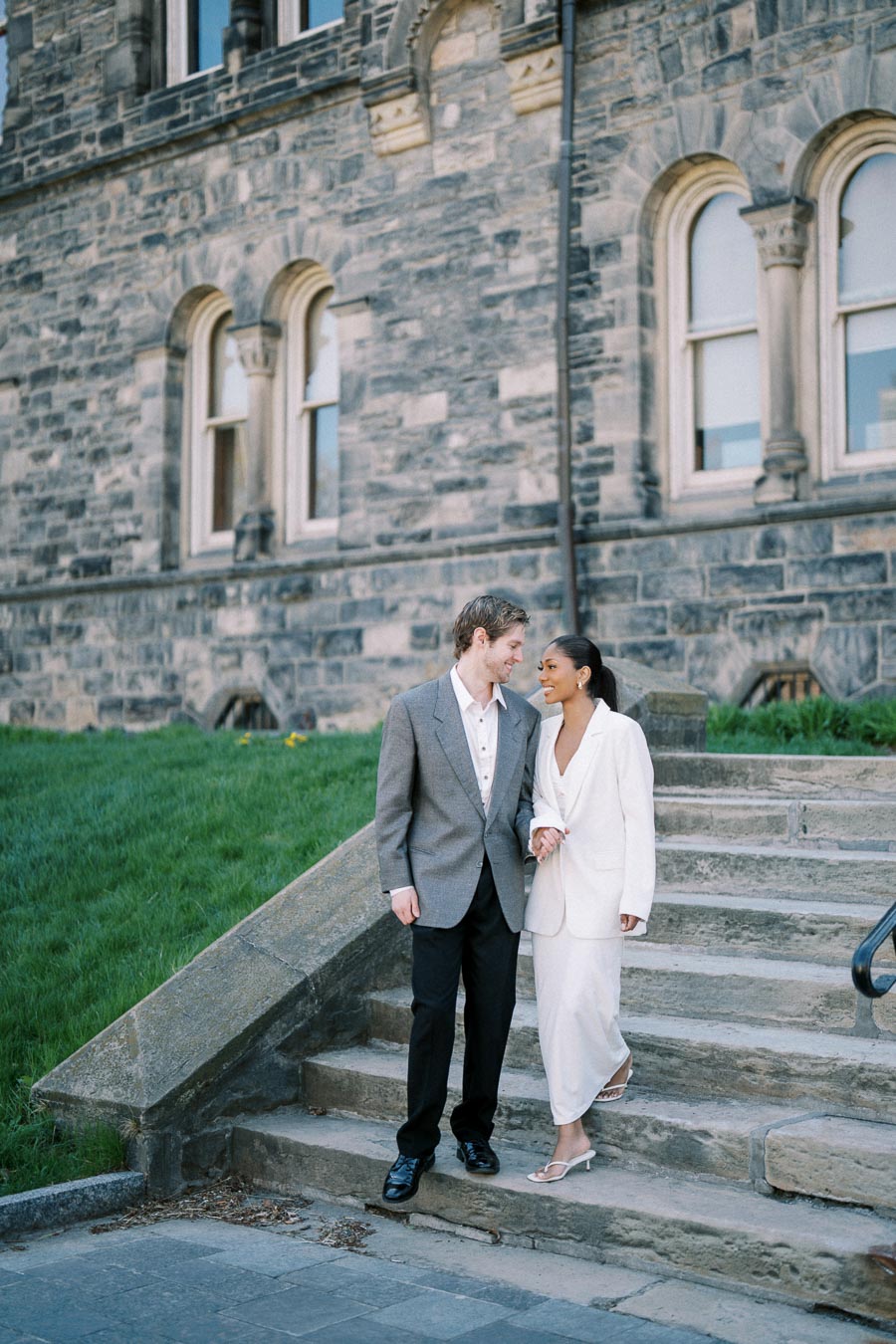 Couple in formal attire smiling on stone steps with historic brick building backdrop.