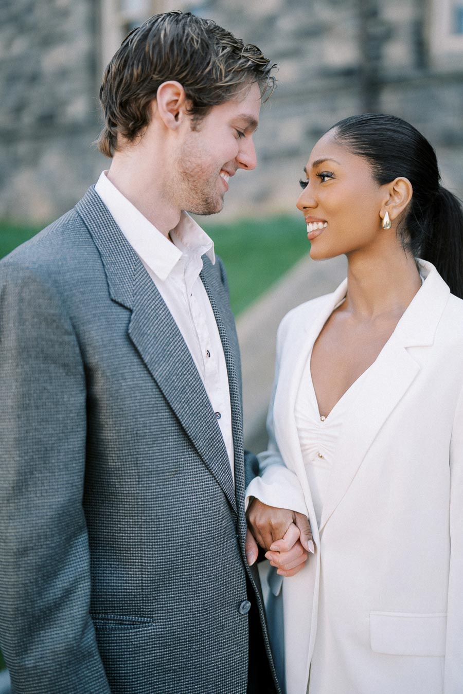Couple in elegant outfits smiling at each other, holding hands, standing outdoors.