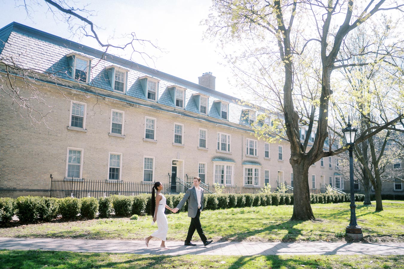 Couple walking hand in hand on a sunny day next to a historic building and trees.