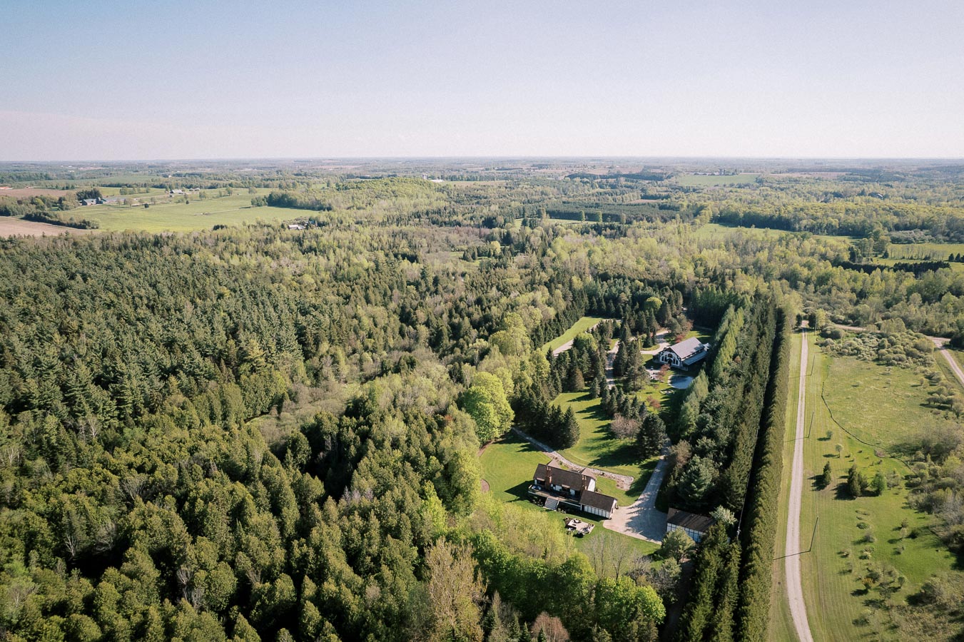 Aerial view of a lush green landscape with dense forests, open fields, and rural houses. The image captures a serene countryside setting with expansive greenery under a clear blue sky.