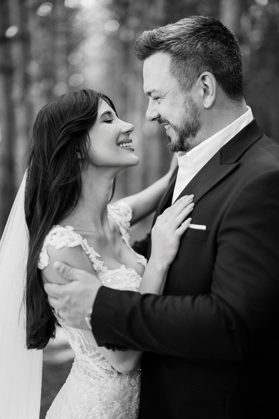 Black and white photo of a joyful bride and groom embracing on their wedding day, surrounded by a serene forest backdrop.