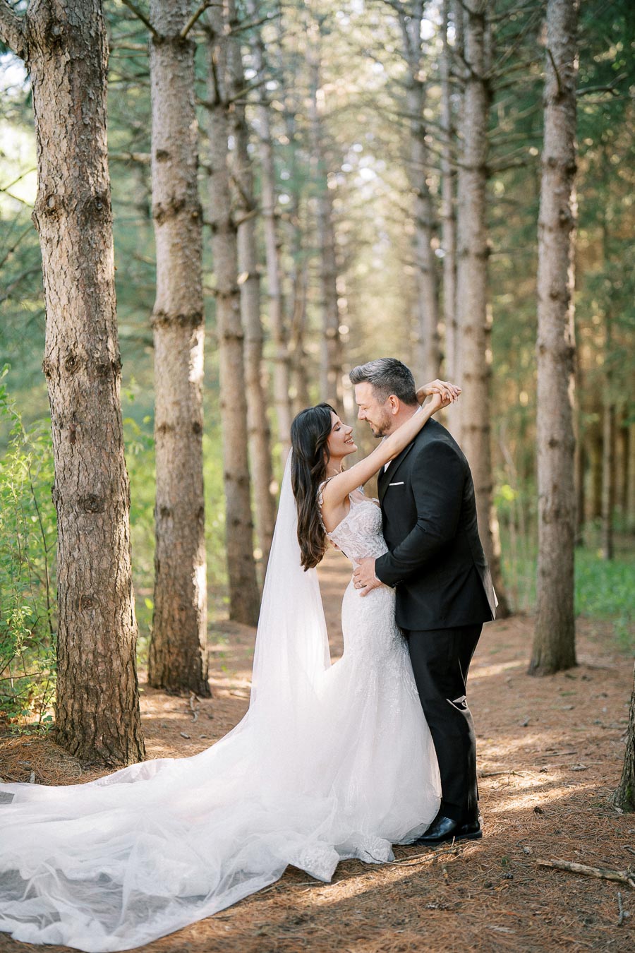 Romantic wedding couple embracing in a serene forest setting, sunlight filtering through the trees, bride in elegant white gown with long train and groom in classic black suit.