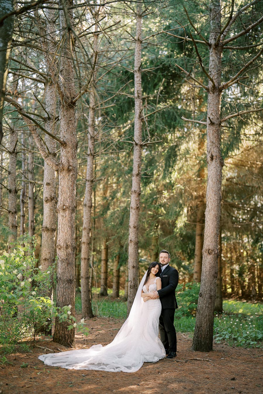 A bride in a flowing white gown and a groom in a black suit embrace in a serene forest setting, surrounded by tall trees and dappled sunlight filtering through the branches.