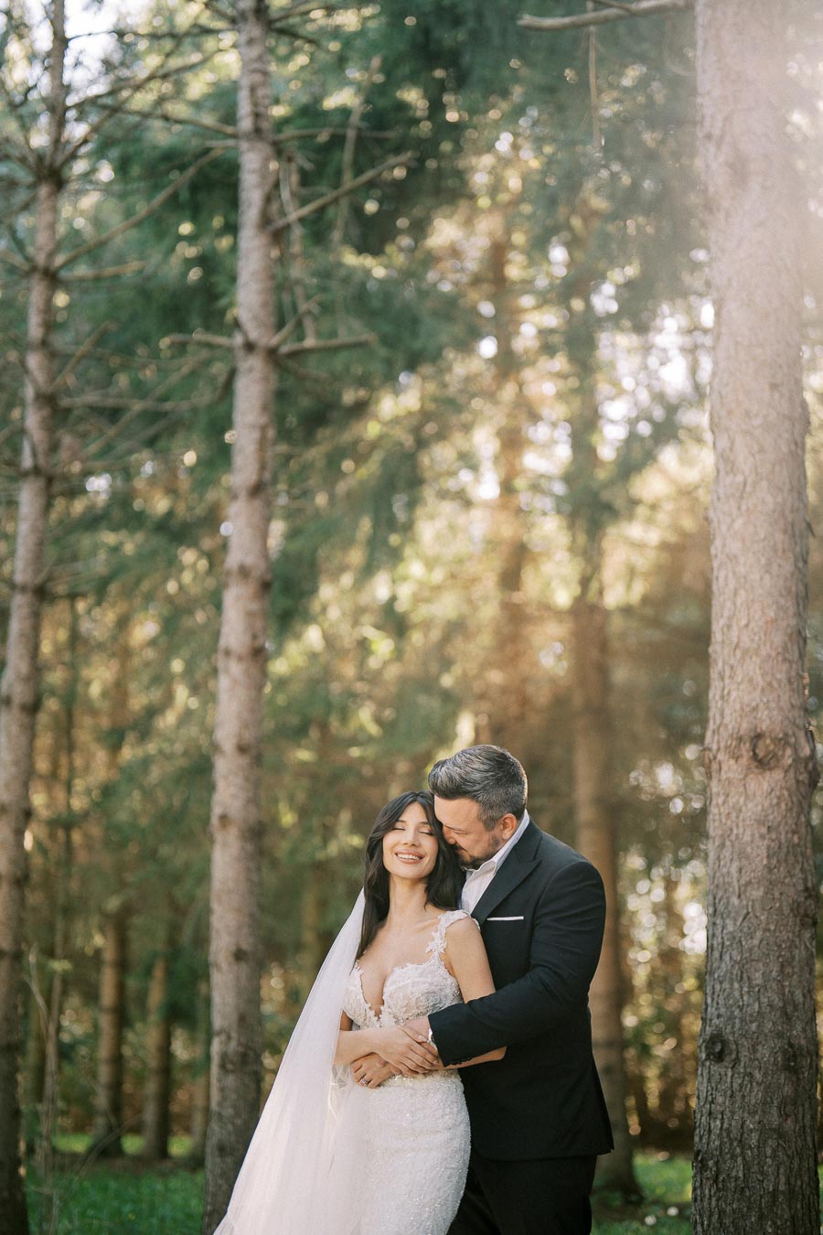 A newlywed couple embraces in a sunlit forest setting, with the bride in a lace wedding gown and the groom in a black suit, surrounded by tall trees and dappled sunlight.
