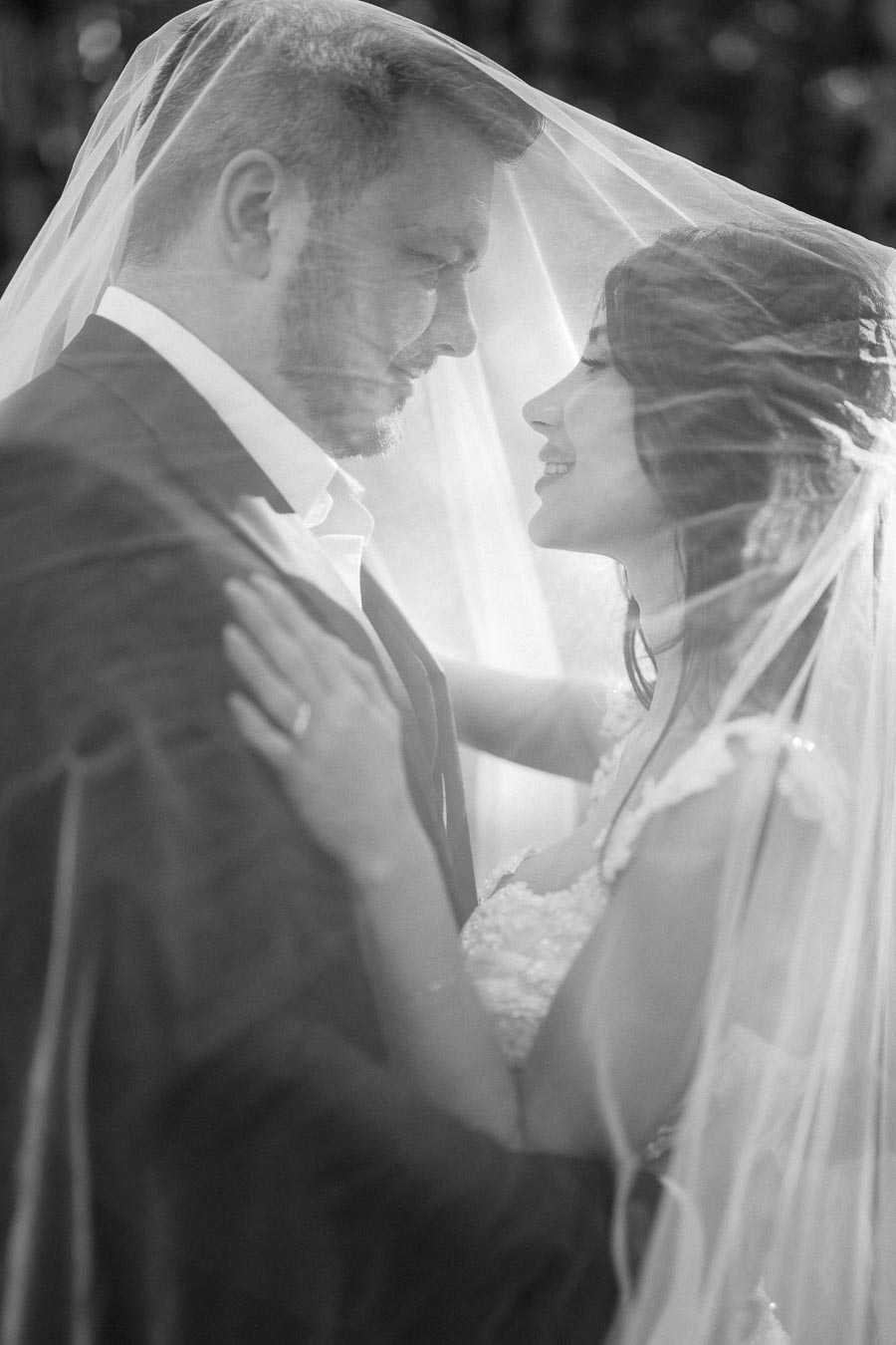 A black and white image of a bride and groom embracing intimately under a sheer veil, capturing a tender wedding moment.