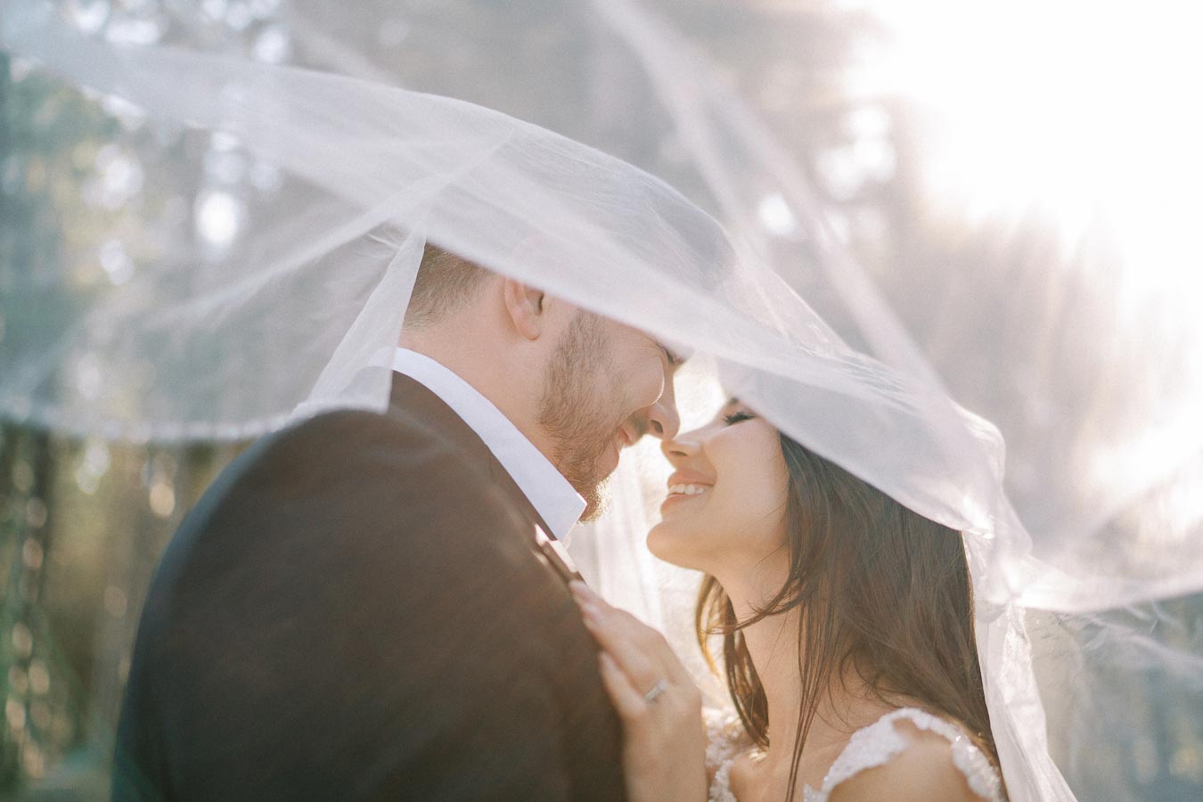 Romantic wedding couple sharing an intimate moment under a veil, with sunlight filtering through trees in the background.