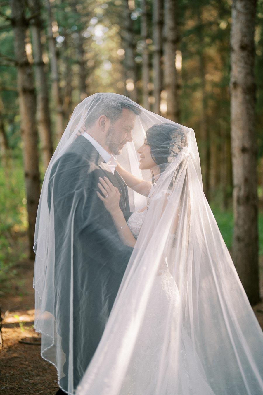 A bride and groom sharing a romantic moment under a veil in a forest setting, with sunlight filtering through the trees.