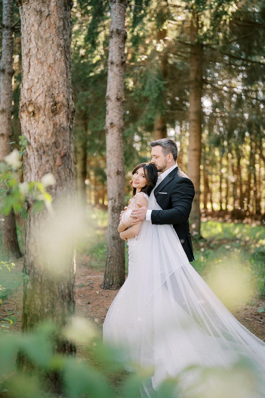 A bride in a flowing white wedding gown and a groom in a stylish black suit sharing an intimate moment in a sunlit forest setting.