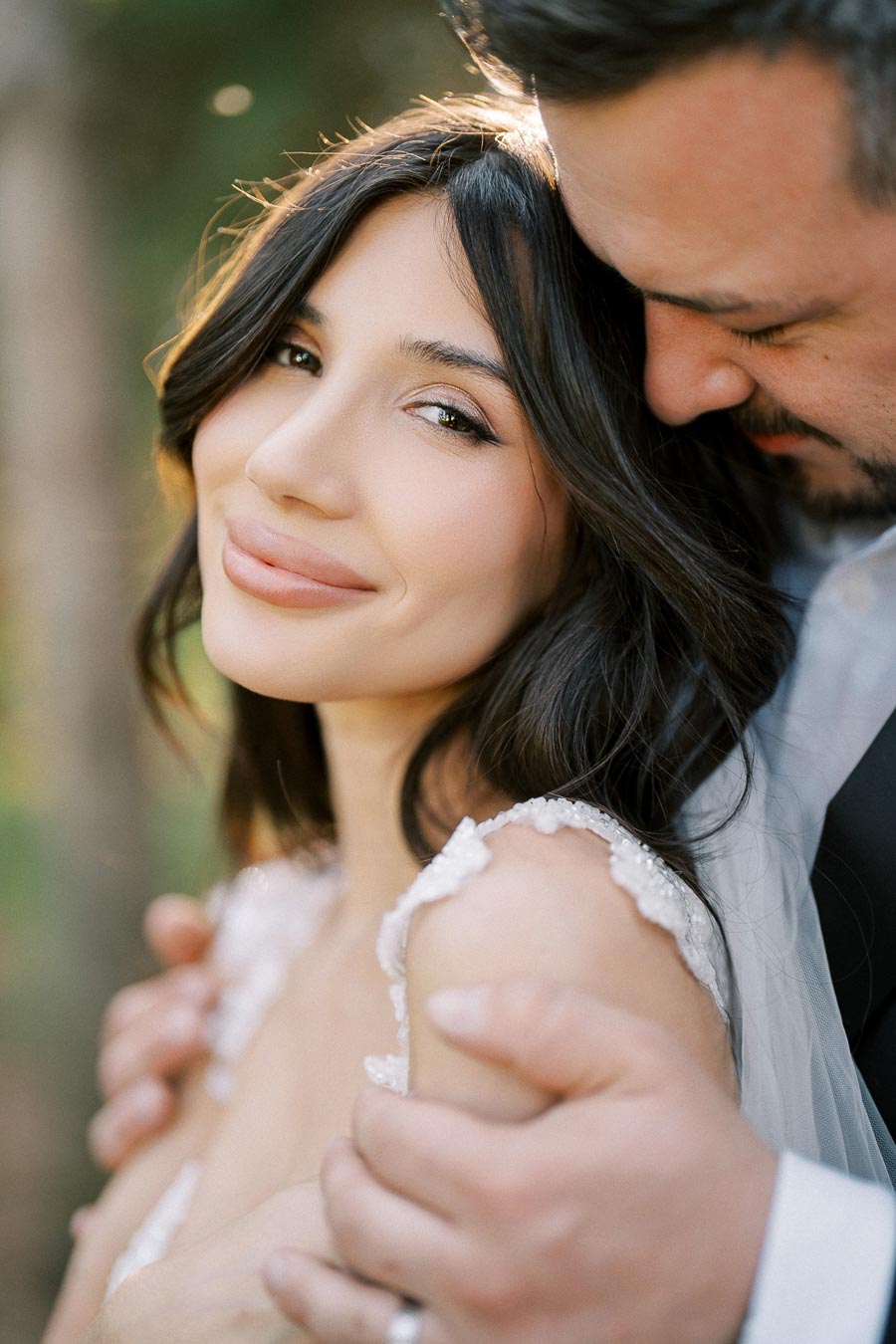 A romantic portrait of a couple on their wedding day, featuring the bride in an elegant gown with delicate lace detailing, smiling softly as her partner embraces her gently, set against a natural outdoor background.