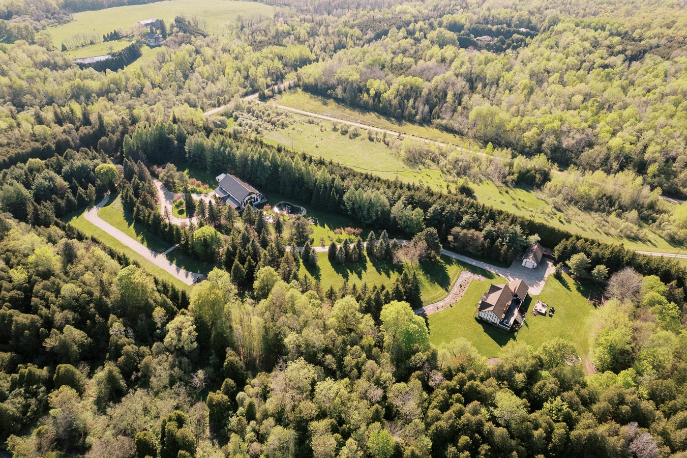 Aerial view of a countryside property surrounded by lush green trees and fields, featuring two large houses with winding driveways, set amidst a vibrant natural landscape.