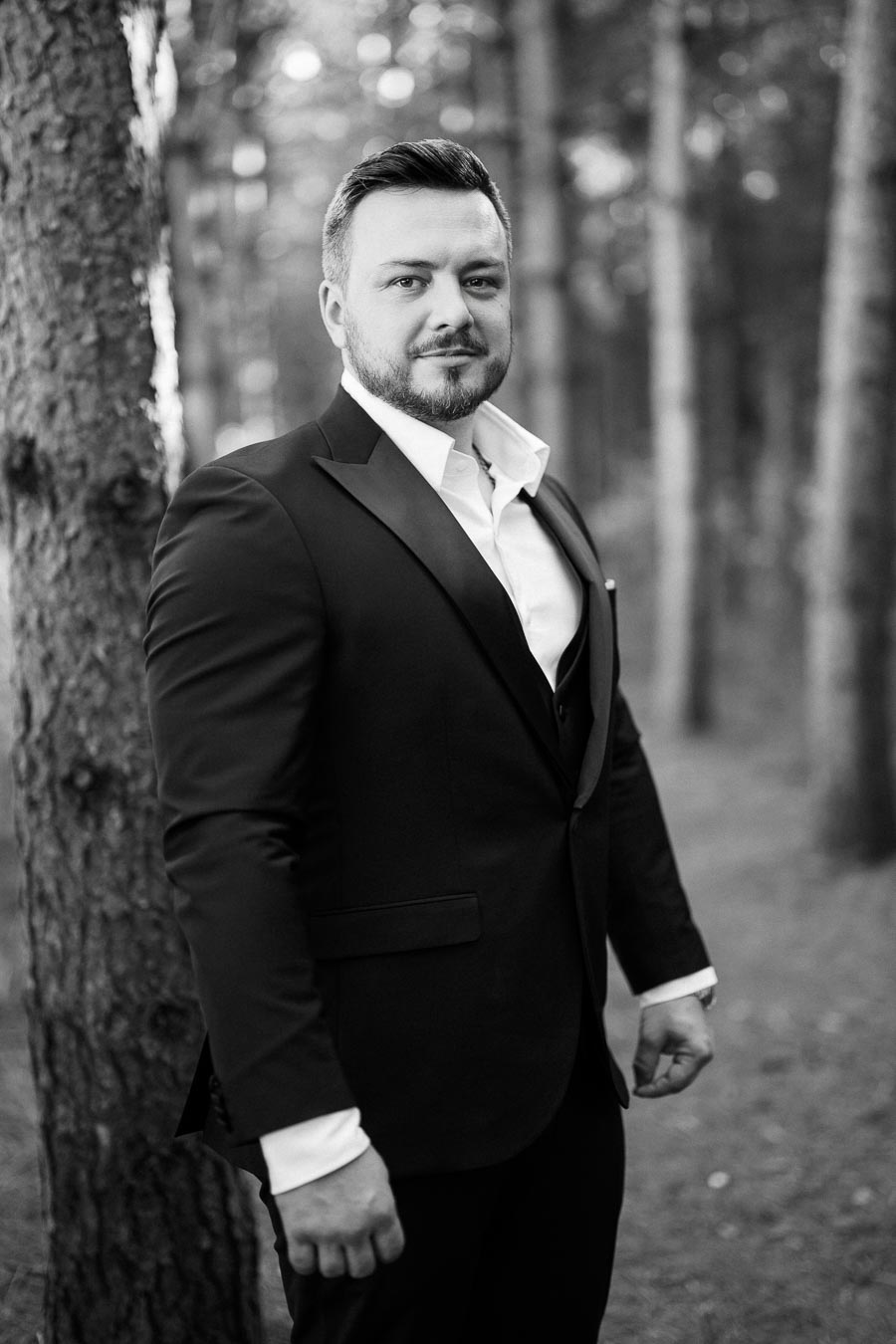 A man in a formal suit standing in a forest, exuding confidence and elegance. Black and white portrait photography.