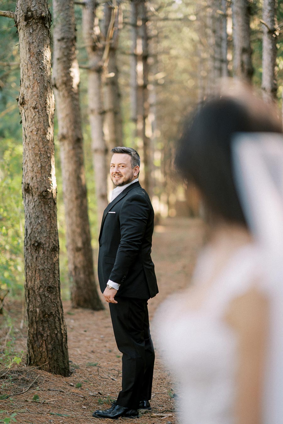 Man in a suit smiling among trees in a forest setting, with an out-of-focus figure in the foreground.