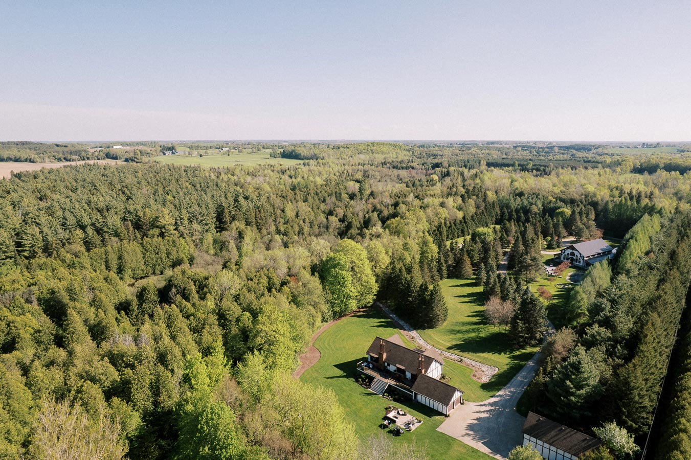 Aerial view of a vast green landscape with dense forest and scattered farmhouses under a clear blue sky.