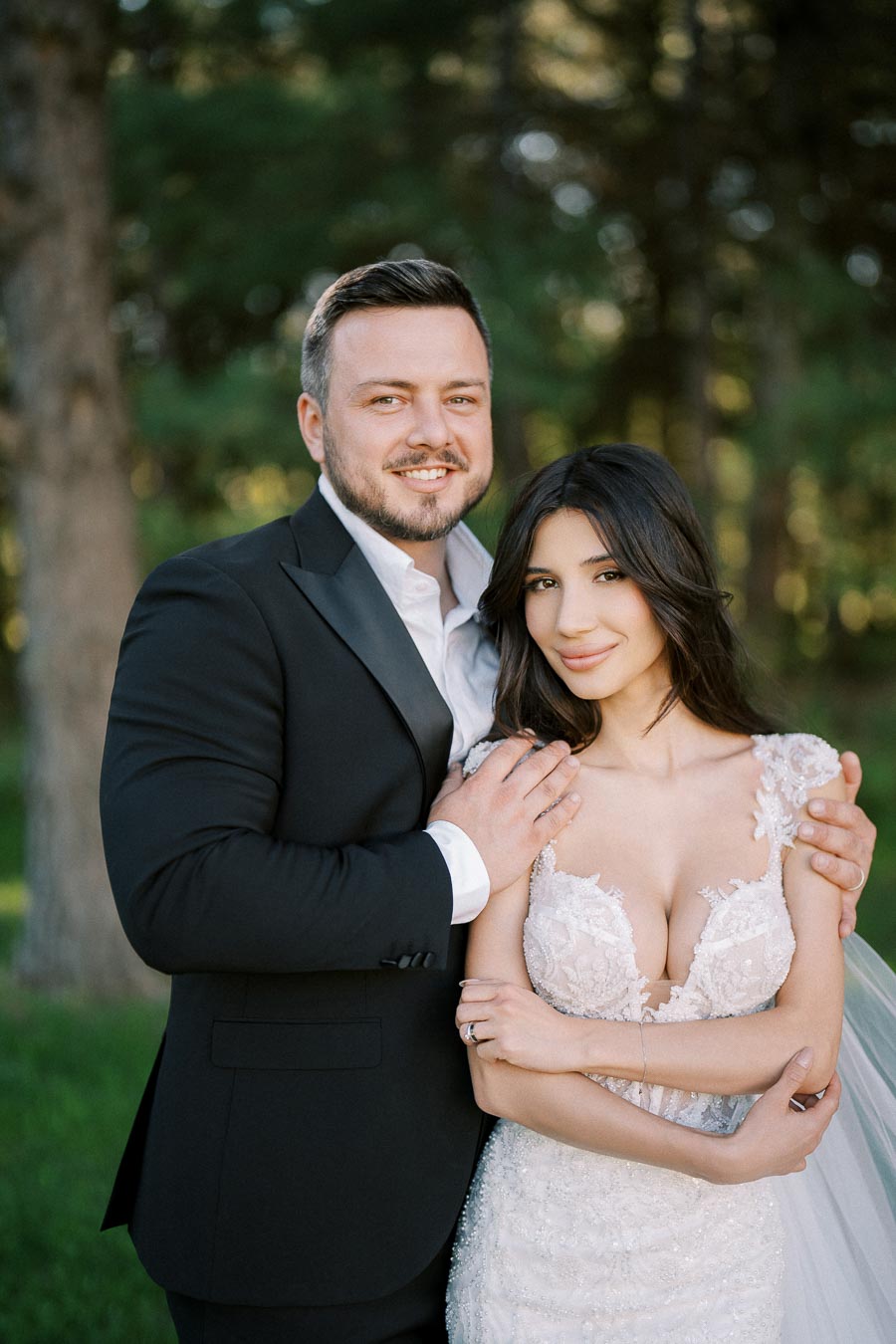 A bride and groom in wedding attire smiling in an outdoor setting with trees in the background.
