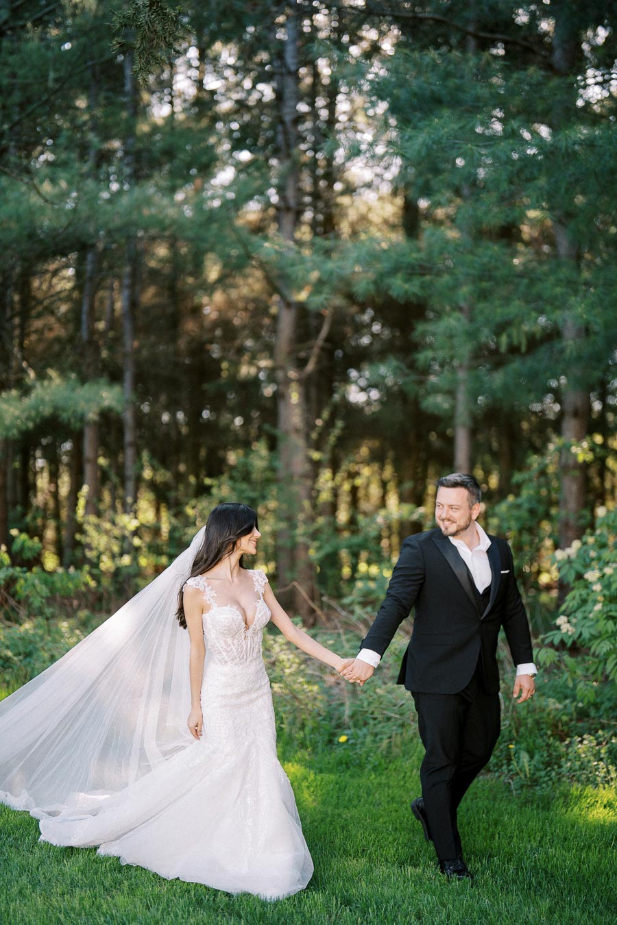 A bride in a flowing white wedding dress and veil holds hands with a groom in a black suit, walking through a lush green forest setting.
