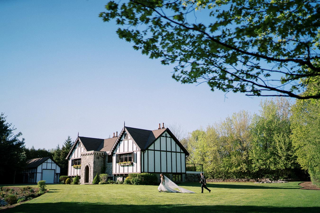A bride and groom walk across a sunny, lush green lawn towards a charming Tudor-style house, surrounded by vibrant trees and a clear blue sky.