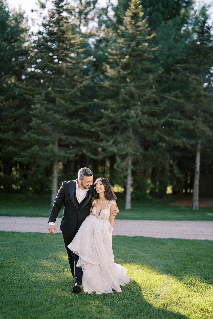 A happy newlywed couple walking arm in arm on a grassy path with a backdrop of lush green trees, the bride in a flowing white gown and the groom in a black suit, both smiling joyfully.