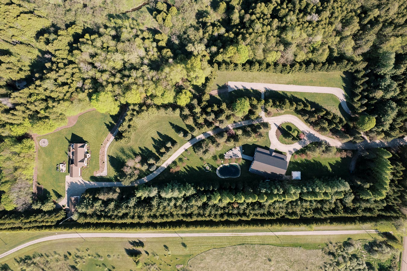 Aerial view of a rural landscape featuring a large house surrounded by lush greenery, winding driveways, and dense woods, showcasing a tranquil natural setting.