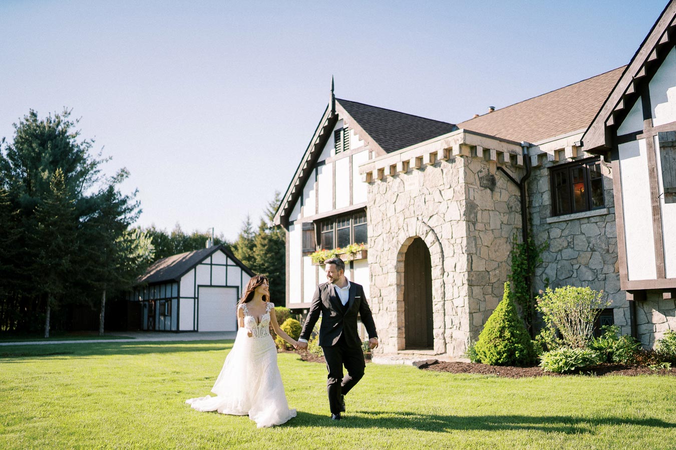 A bride and groom holding hands and walking on a grassy lawn in front of a large stone building with a rustic, Tudor-style design on a sunny day, surrounded by lush greenery.