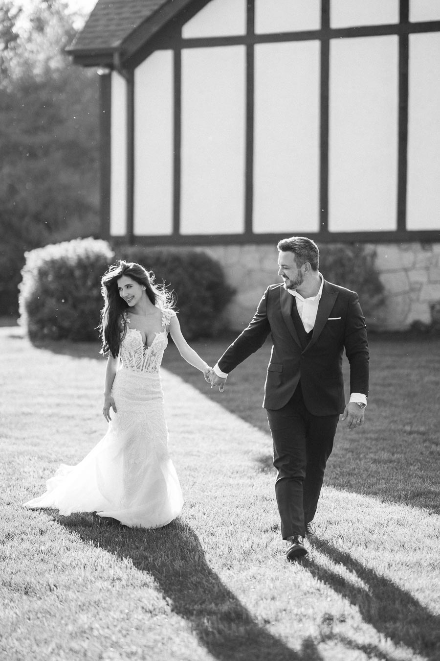 Black and white photo of a joyful bride and groom holding hands while walking on a sunny lawn, with a quaint building in the background, capturing a serene outdoor wedding moment.
