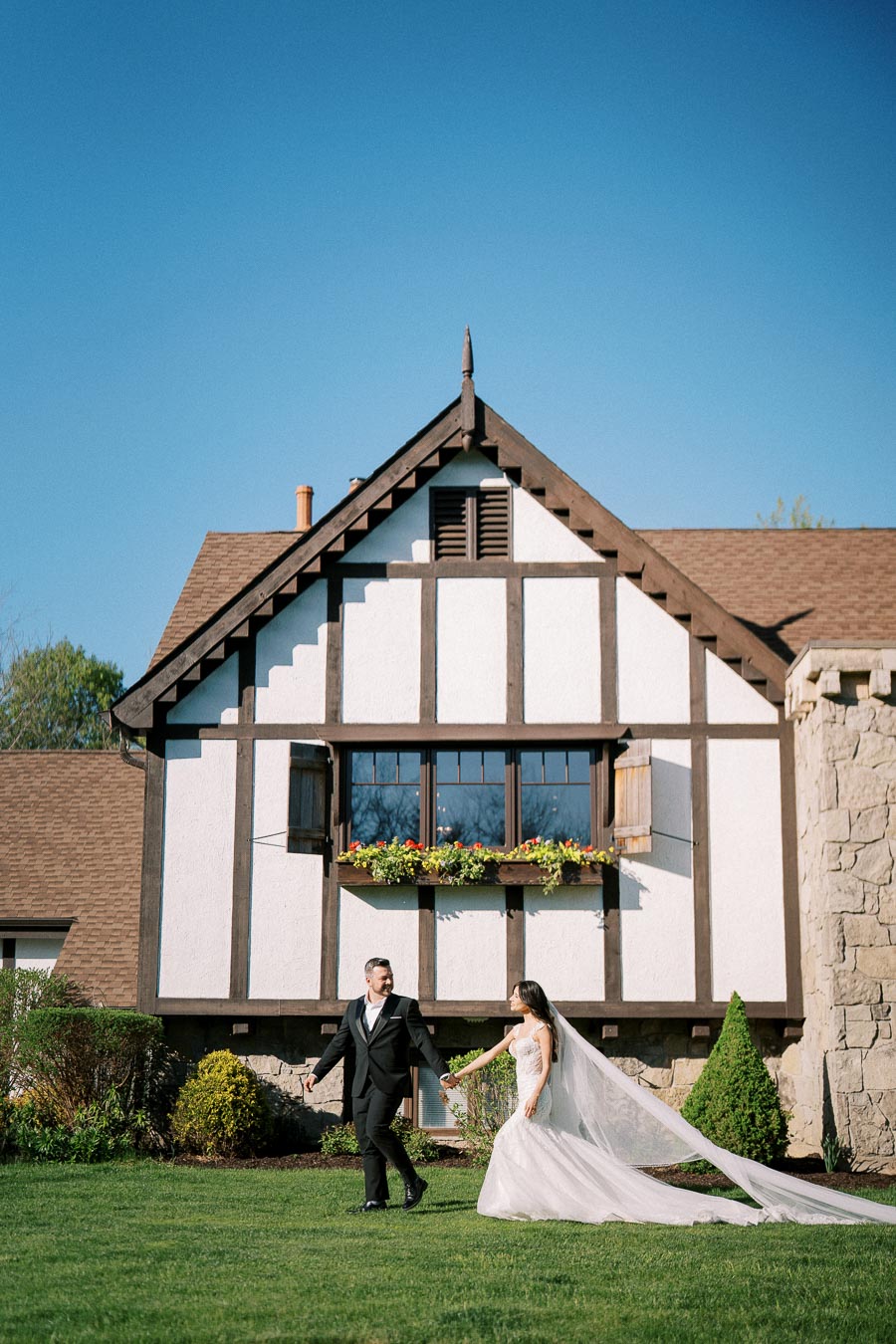 A bride and groom holding hands and smiling while walking on a manicured lawn in front of a charming Tudor-style house, with a vibrant blue sky above.