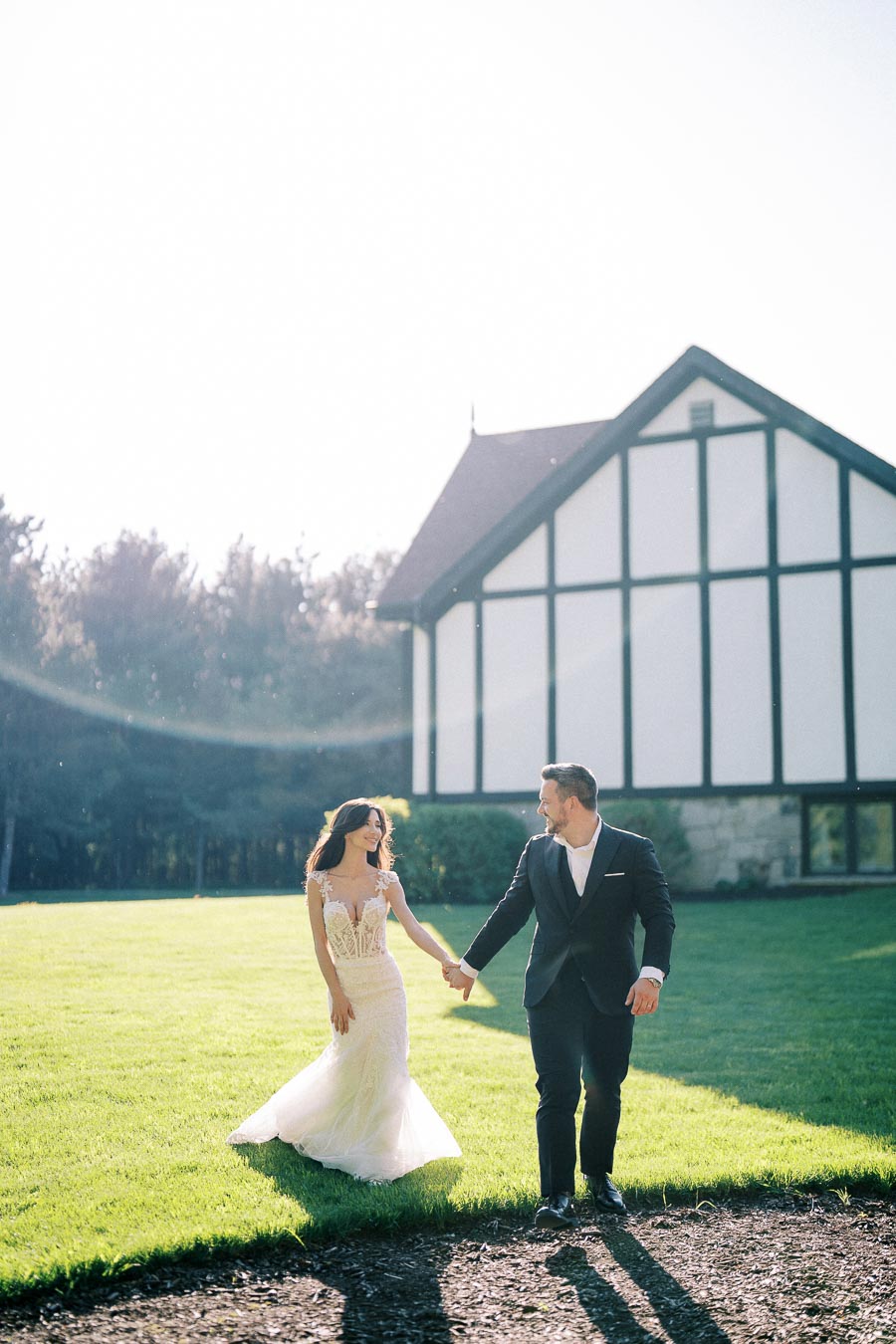A bride in a lace wedding dress and a groom in a dark suit holding hands and walking on a sunlit lawn in front of a white and black Tudor-style building.
