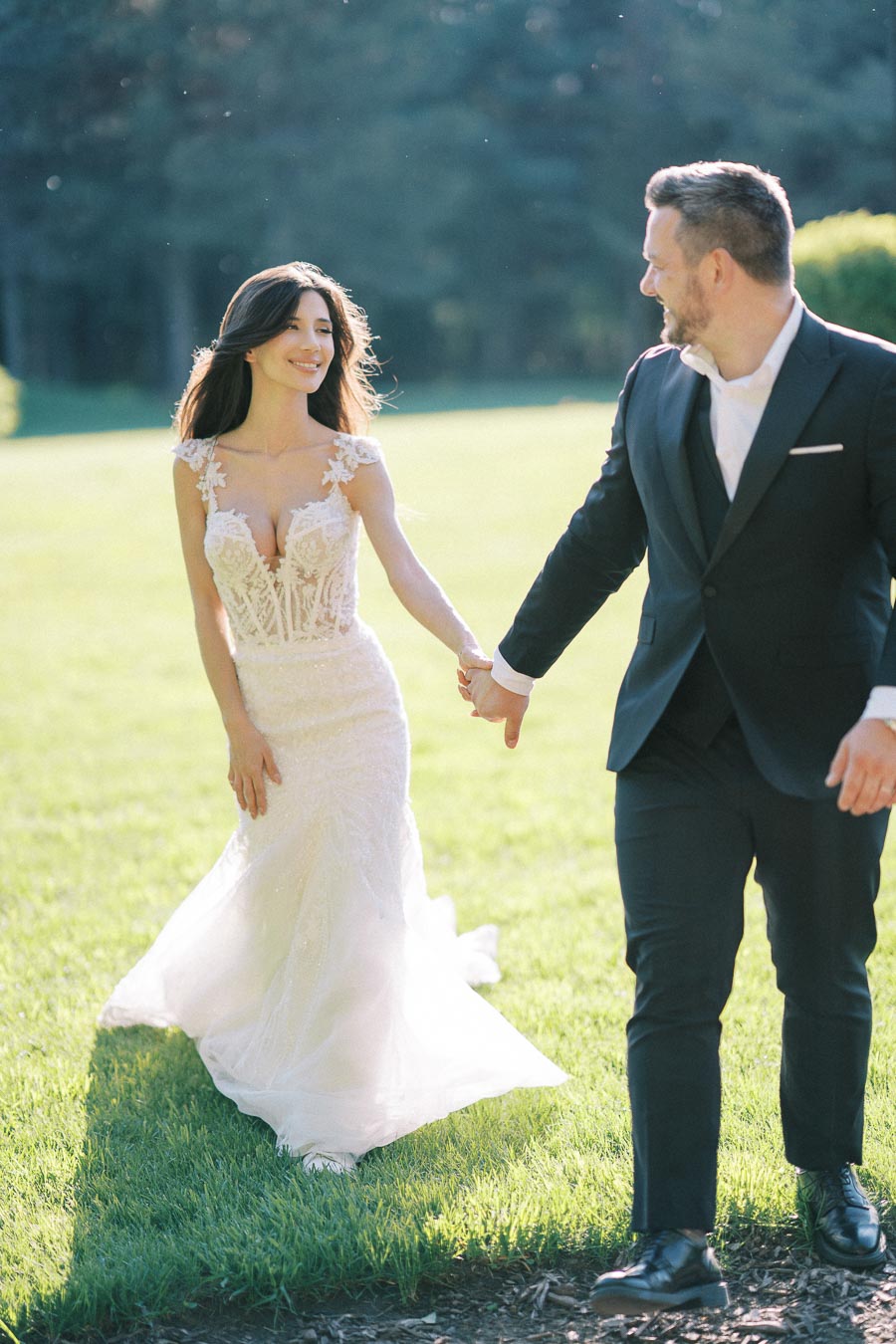 A bride in a lace wedding dress and a groom in a dark suit holding hands while walking on a sunlit grassy field.