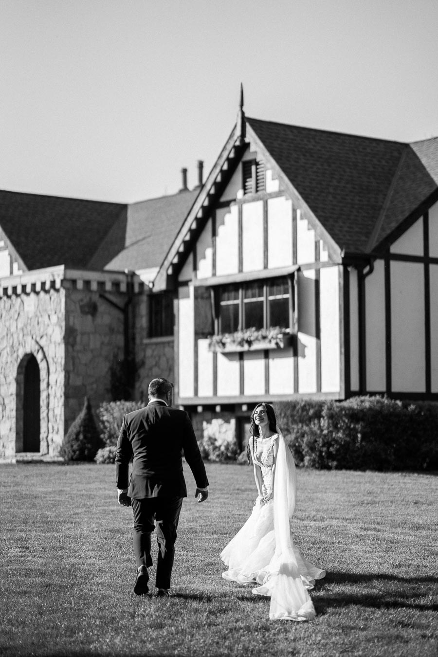 Black and white image of a smiling bride and groom walking towards each other on a lawn in front of a Tudor-style building.