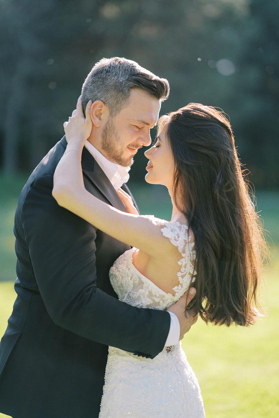 Romantic couple embracing outdoors in elegant wedding attire, bride in lace dress and groom in black suit, captured in a sunlit garden setting.
