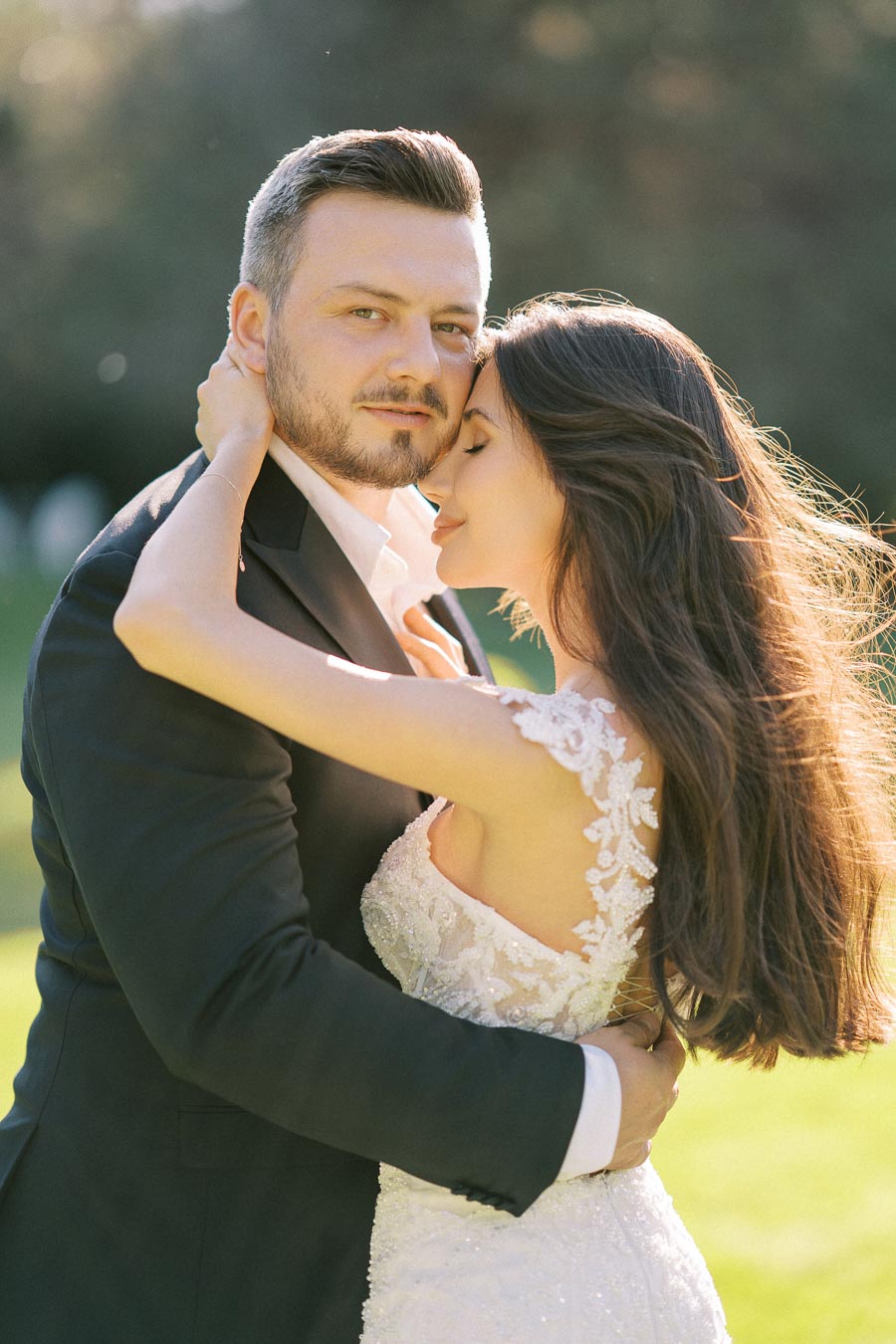 A couple embraces lovingly in a sunlit garden, with the woman wearing an elegant lace wedding dress and the man in a tailored black suit, capturing a romantic moment.