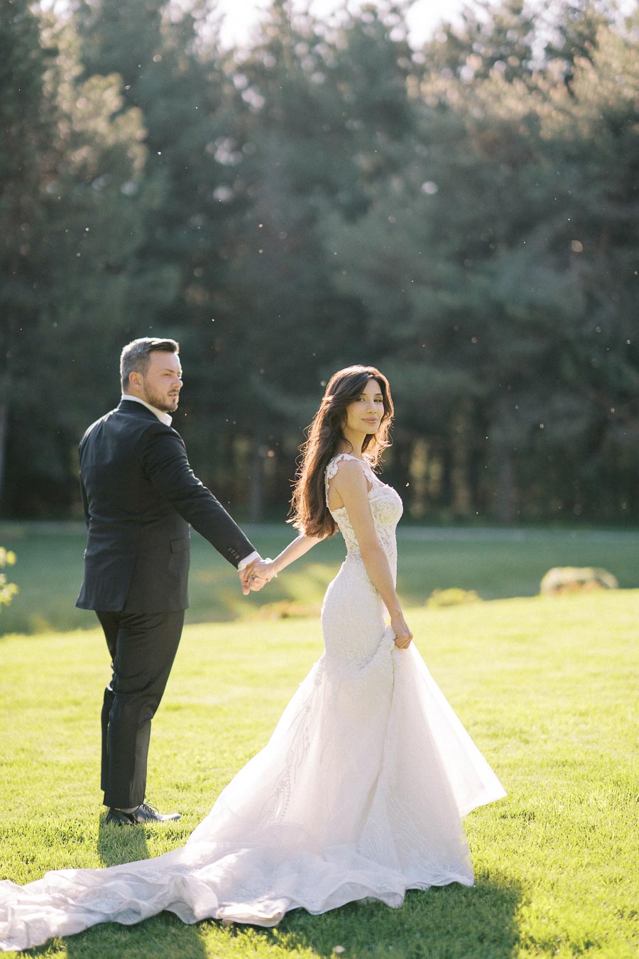 A bride and groom holding hands in a sunlit garden, with the bride wearing a lace wedding gown and the groom in a black suit.