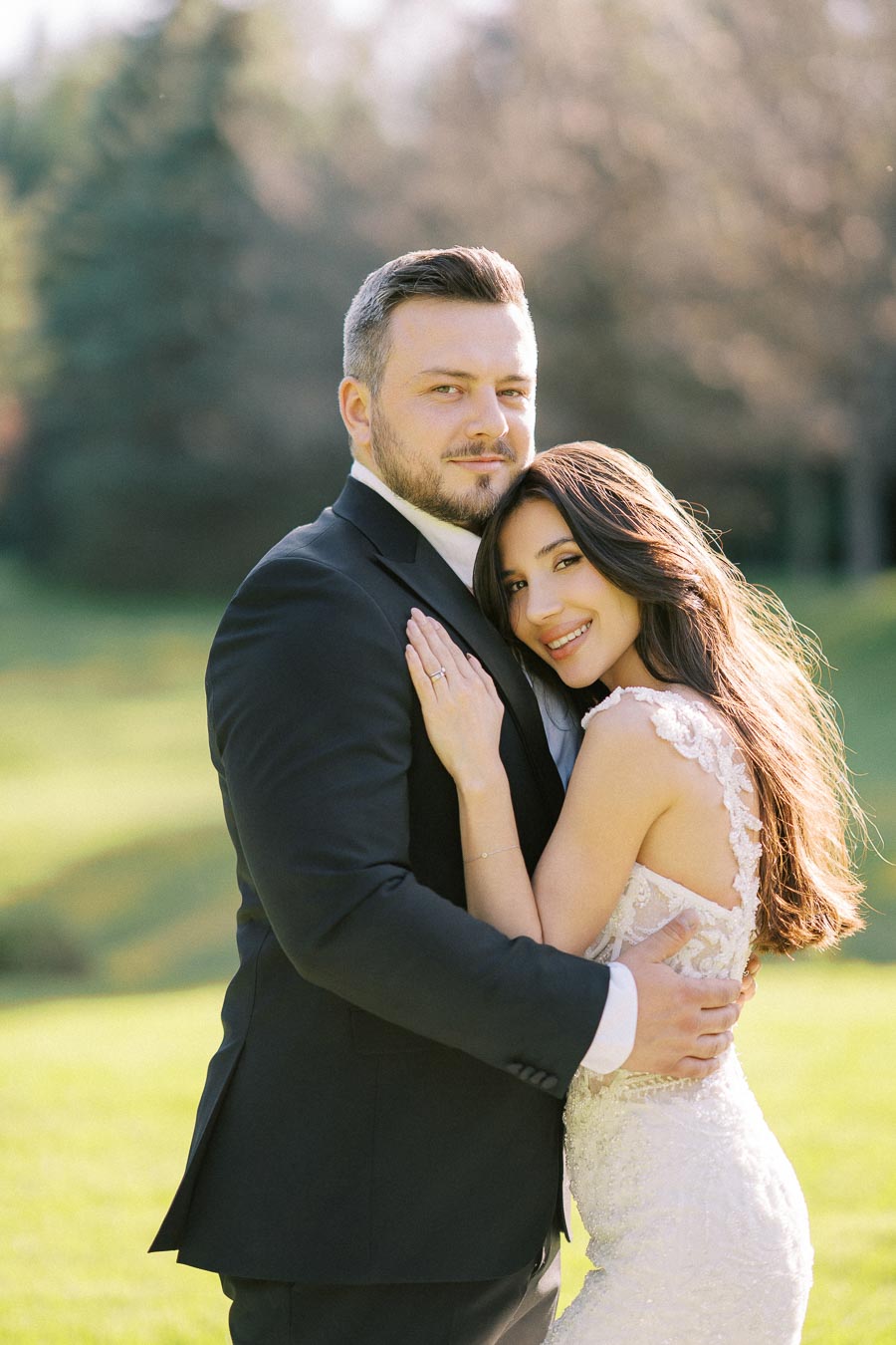 A happy couple in wedding attire embracing outdoors on a sunny day, with green trees and grass in the background.