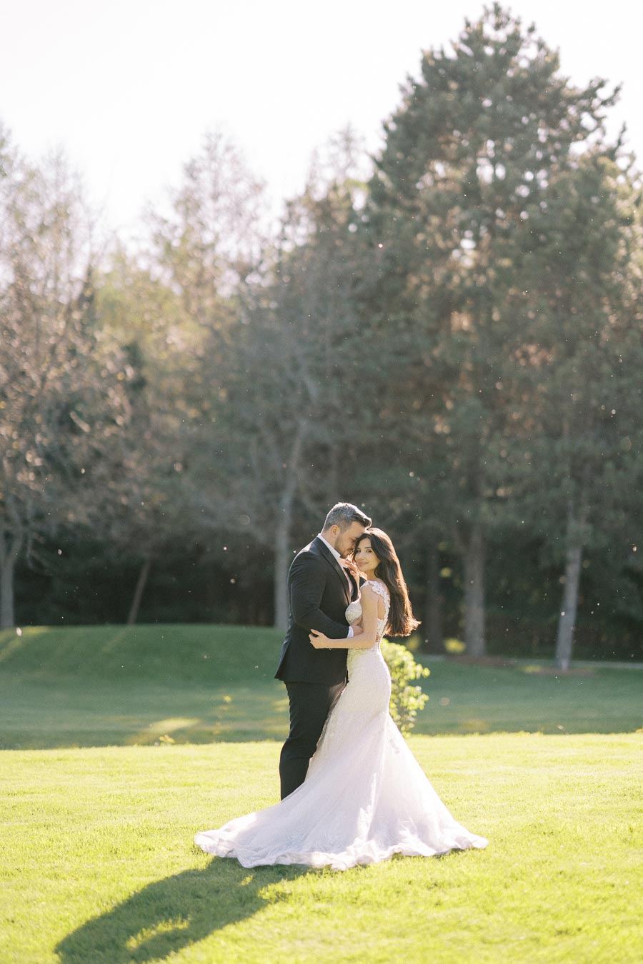 A bride and groom embrace on a sunny day, standing on a lush green lawn with trees in the background, captured in soft, romantic lighting.