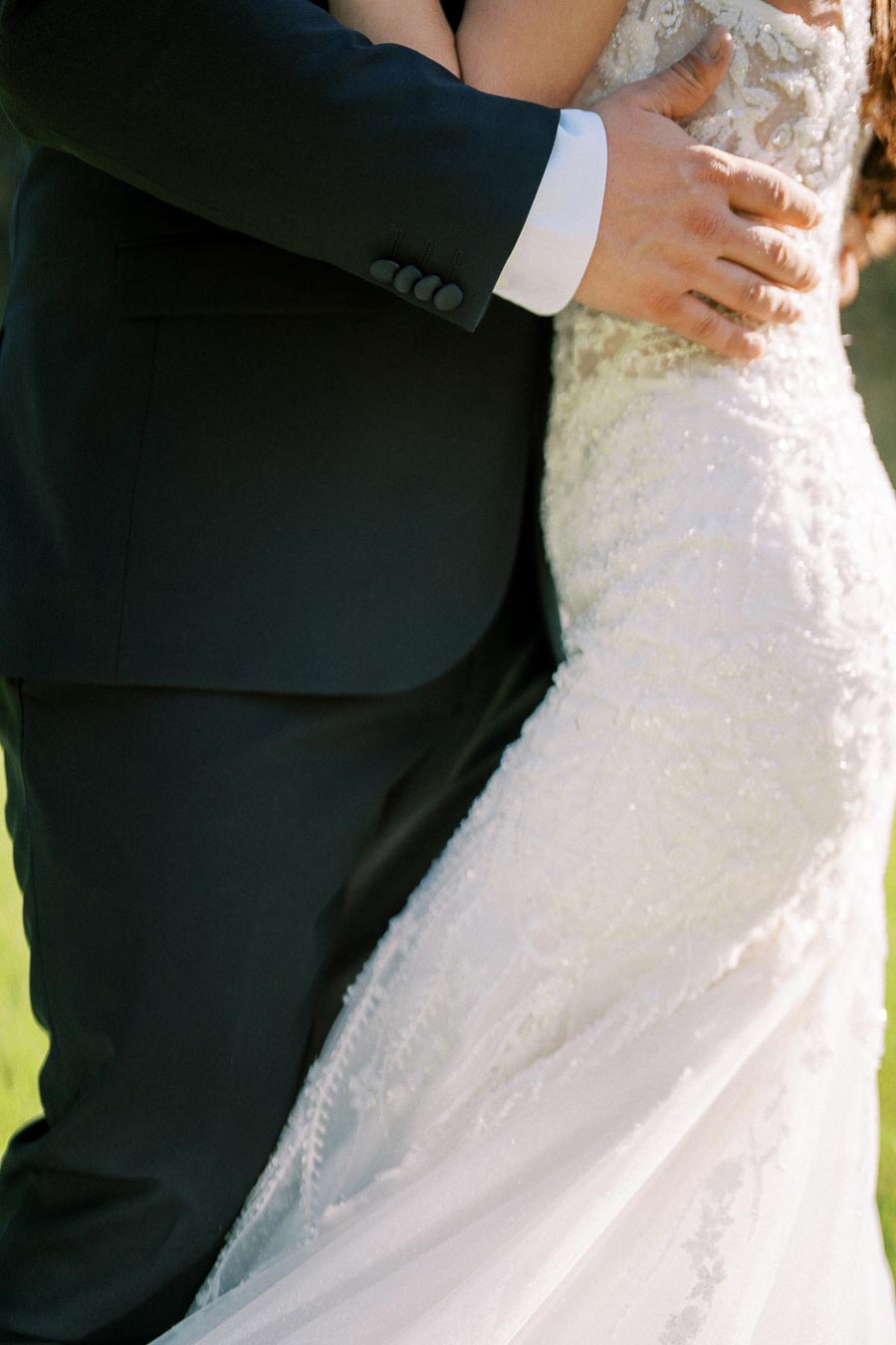 A close-up of a groom in a black suit embracing a bride in a detailed lace wedding gown, capturing an intimate moment on their wedding day outdoors.