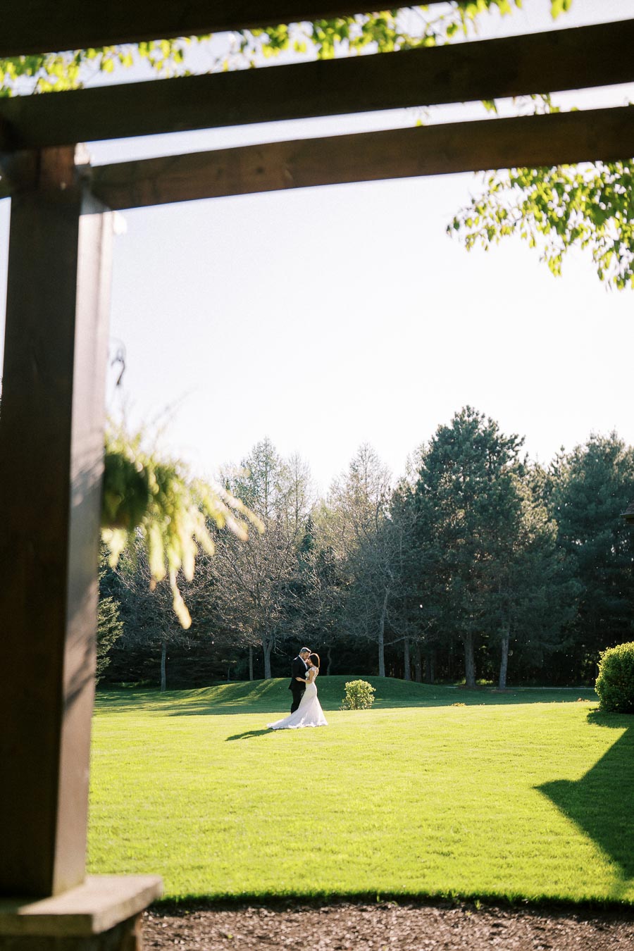 A bride and groom dancing on a sunlit lawn surrounded by trees, captured through a wooden pergola, with greenery in the foreground.
