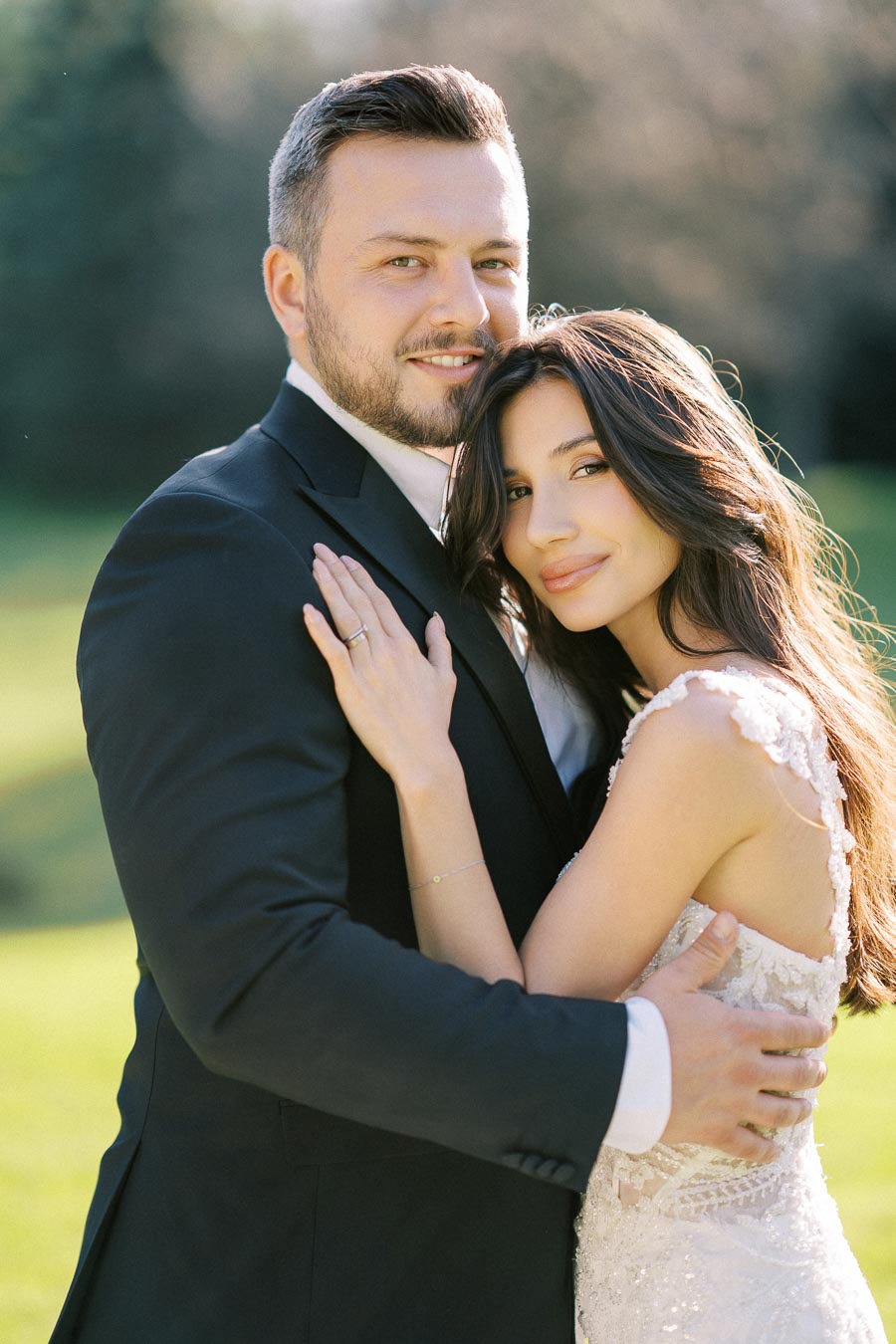 A couple embracing on their wedding day, with the groom in a black suit and the bride in a white lace dress, set against a blurred outdoor background.