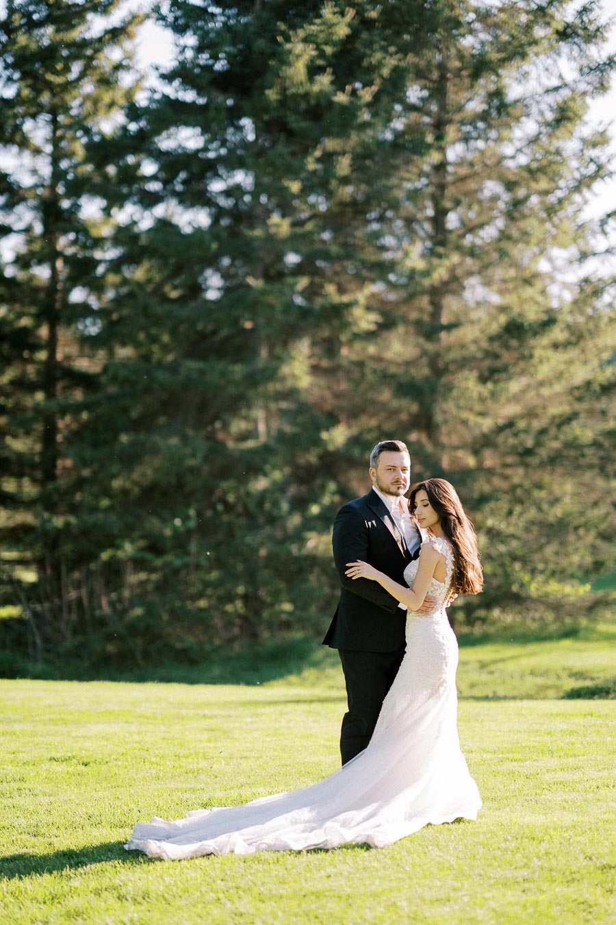Elegant bride and groom embracing in a sunlit garden with lush green trees, symbolizing a romantic outdoor wedding scene.