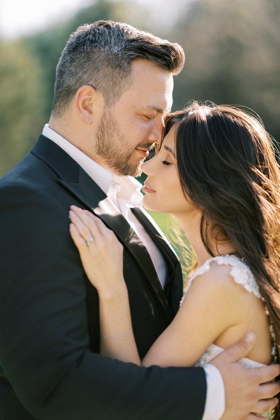 A couple in formal attire embracing outdoors, with the man in a black suit and the woman in a white dress, conveying a sense of romance and affection in a sunlit setting.