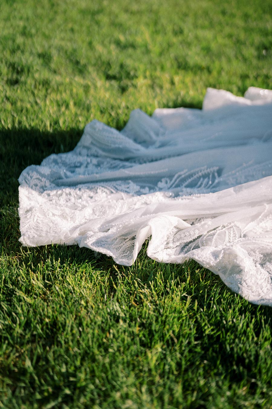 Close-up of a lace wedding dress train draped elegantly on lush green grass, highlighting intricate embroidery and delicate design.