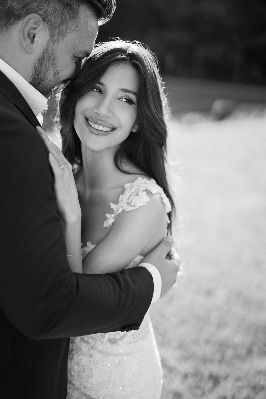 Black and white photo of a happy bride in a lace wedding dress, embraced by a groom in a suit, smiling in a sunlit outdoor setting.