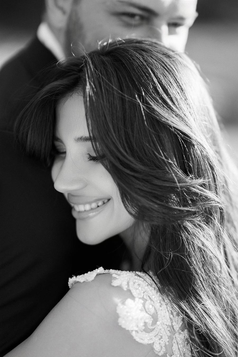 Black and white photo of a woman smiling with her eyes closed, resting against a man's shoulder, wearing a lace wedding dress, conveying a sense of love and happiness.