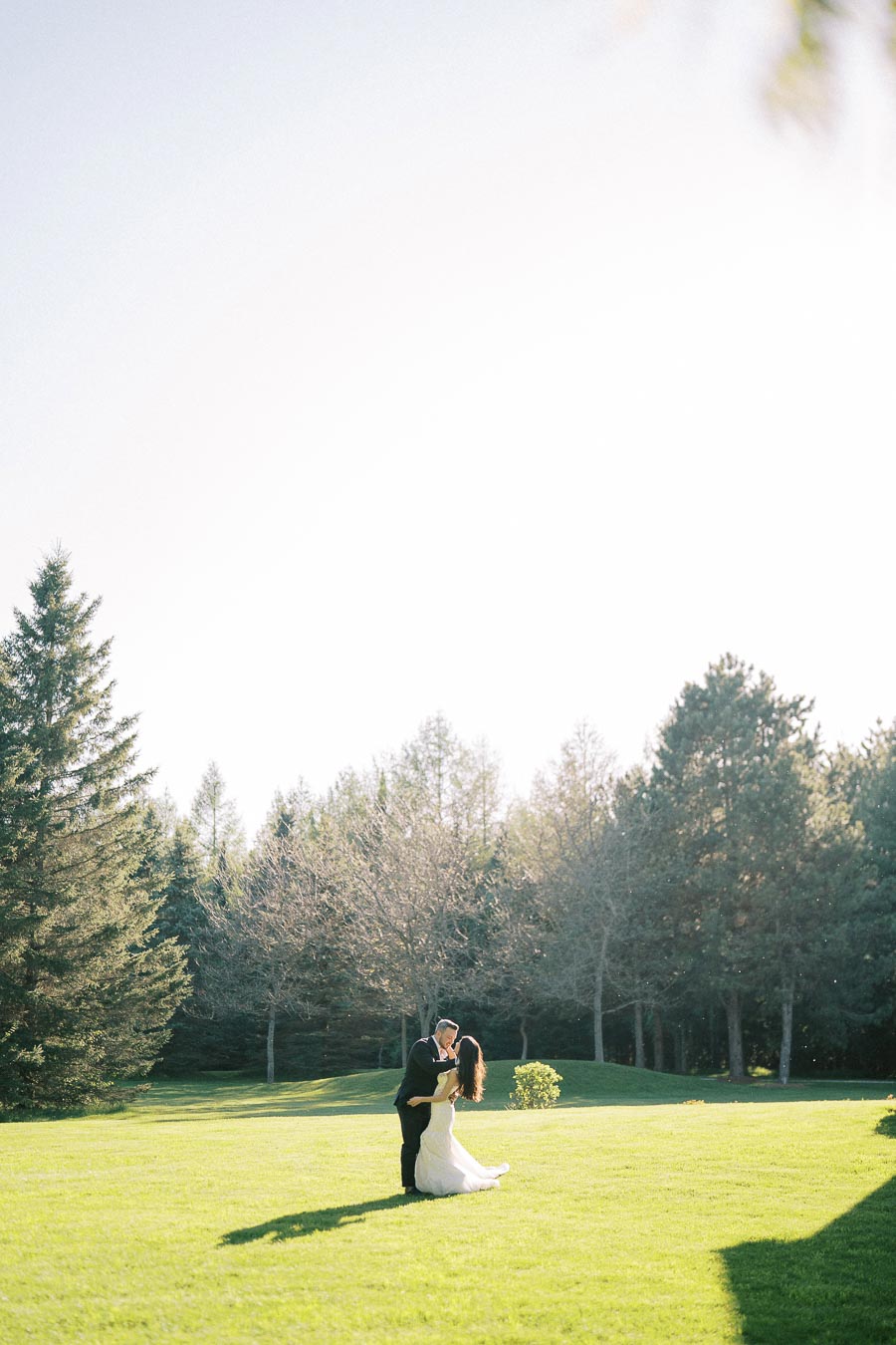 A couple in wedding attire embraces on a sunlit, green lawn surrounded by trees, capturing a romantic outdoor wedding moment.