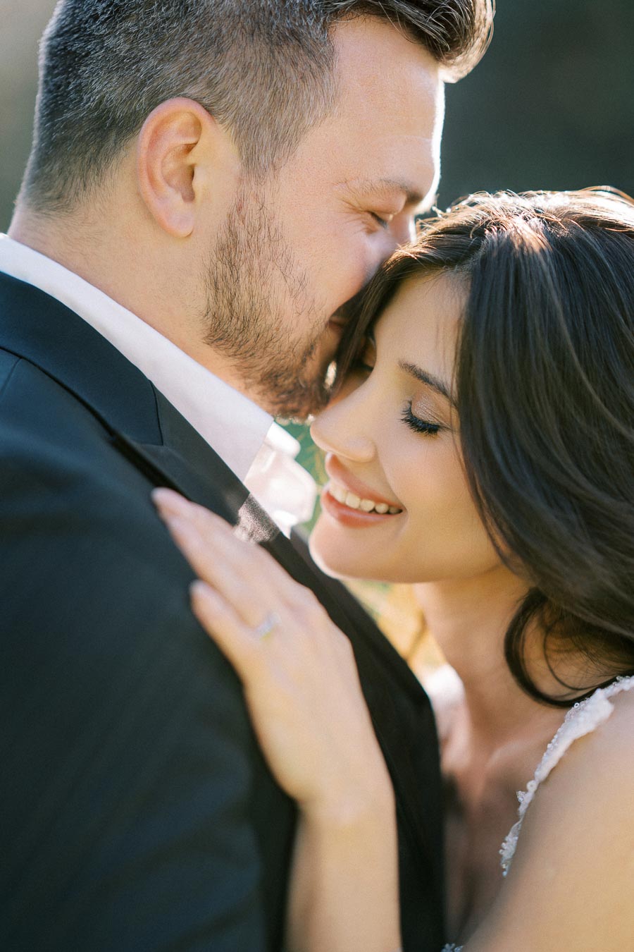 A happy couple embracing closely, with the woman smiling and the man gently kissing her forehead, both dressed elegantly, conveying warmth and affection in a natural outdoor setting.