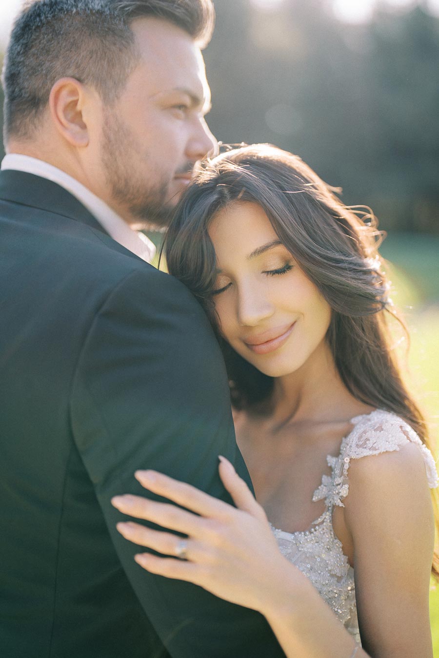 A bride and groom embrace lovingly during a sunny outdoor wedding photo shoot, with the bride resting her head on the groom's shoulder, capturing a tender and romantic moment.